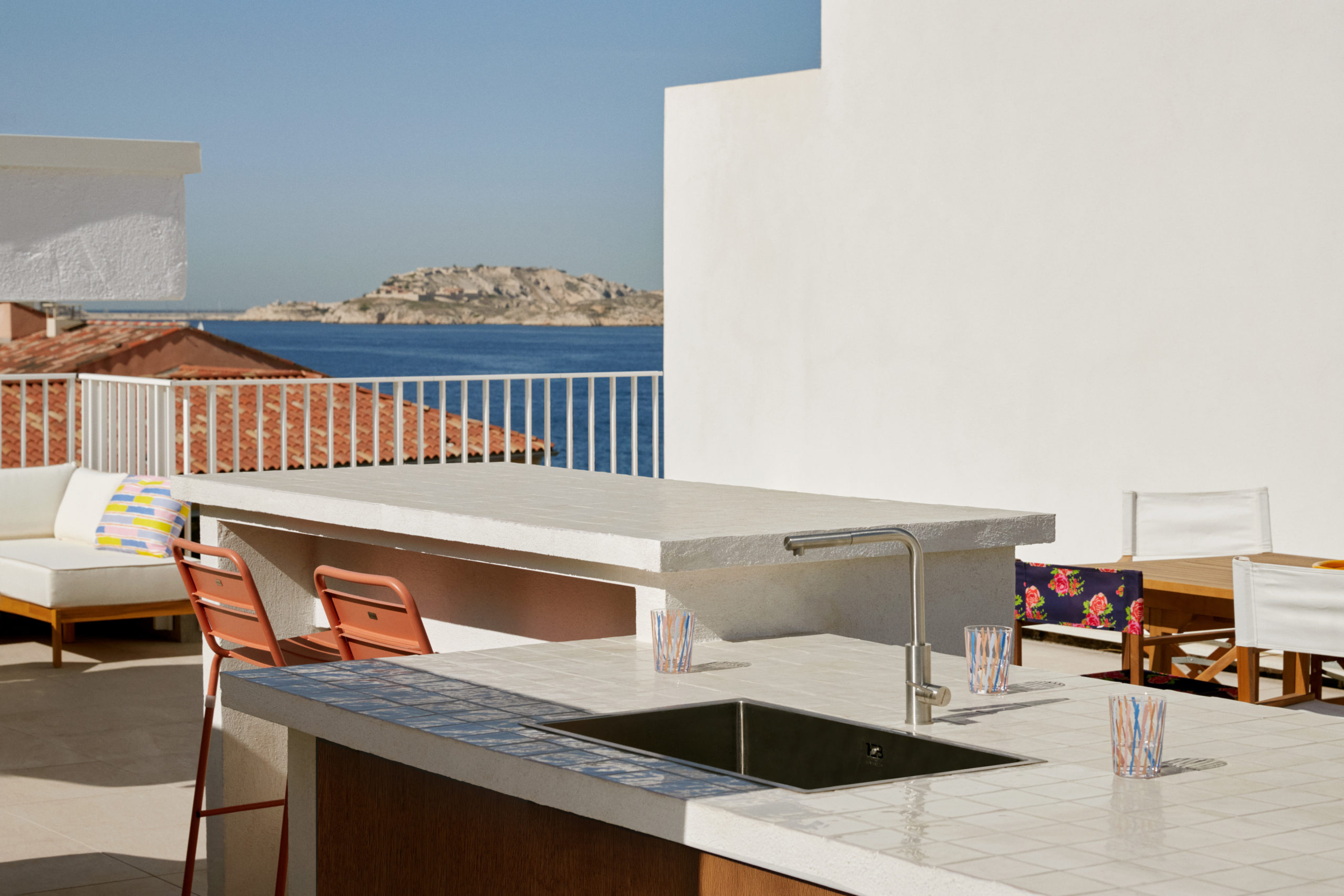 Rénovation d'un appartement et aménagement d'un rooftop - Toit-terrasse avec des dalles sur plot en carrelage Marrazzi - Garde corps en métal blanc barreaudage vertical de la marque Technal - Vue sur la mer et les îles de frioul à Marseille - îlot de cuisine extérieur avec évier et porte en bois - chaise de bar - zellige - chaises plantes motif floral