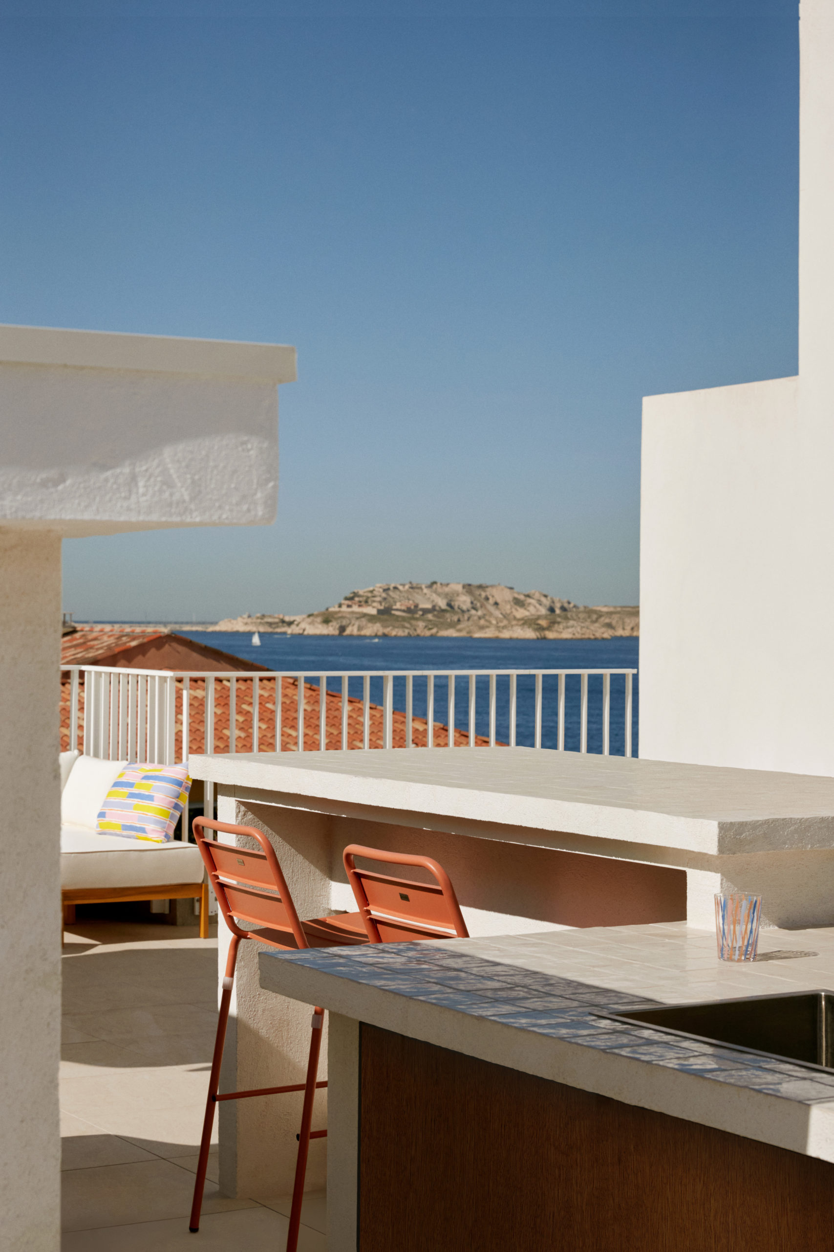 Rooftop - Toit terrasse avec des dalles sur plot en carrelage Marrazzi - Garde corps en métal blanc barreaudage vertical de la marque Technal - Vue sur la mer et les îles de frioul à Marseille - îlot de cuisine extérieur avec évier et porte en bois - chaise de bar - zellige