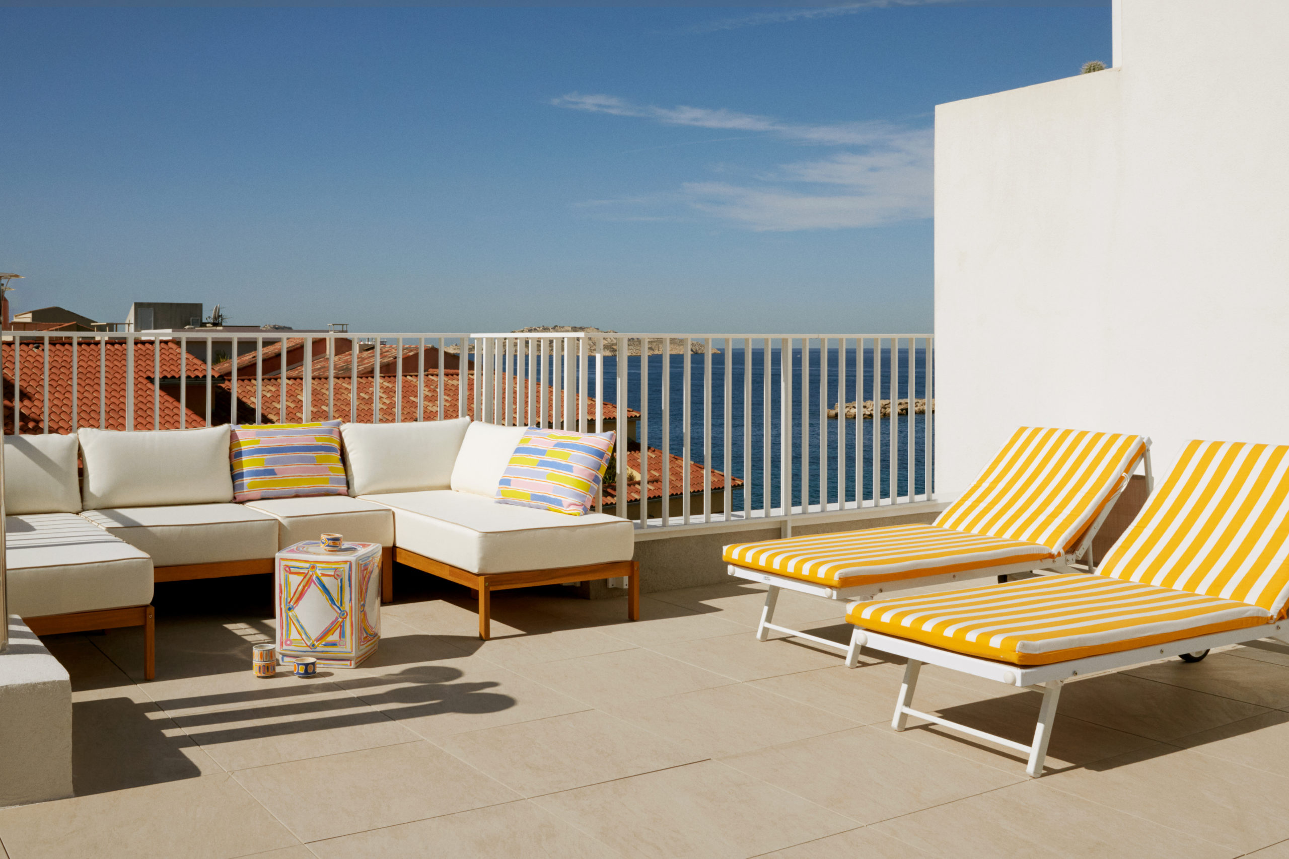 Rénovation d'un appartement et aménagement d'un rooftop - Dalles sur plots en carrelage Marrazzi - Garde corps en métal blanc barreaudage vertical de la marque Technal - Transat avec matelas rayé jaune et blanc - Coin lounge avec ensemble de jardin d'angle en bois - Coussin et table d'appoint collection Designer Monoprix - Vue sur la mer et les îles de frioul à Marseille